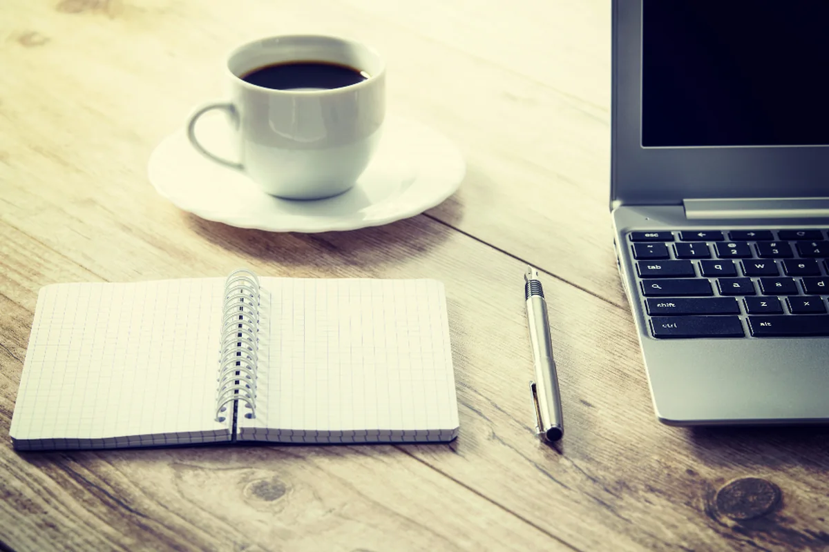Taza de café sobre un platillo, un cuaderno de espiral abierto, un bolígrafo y un portátil sobre una mesa de madera