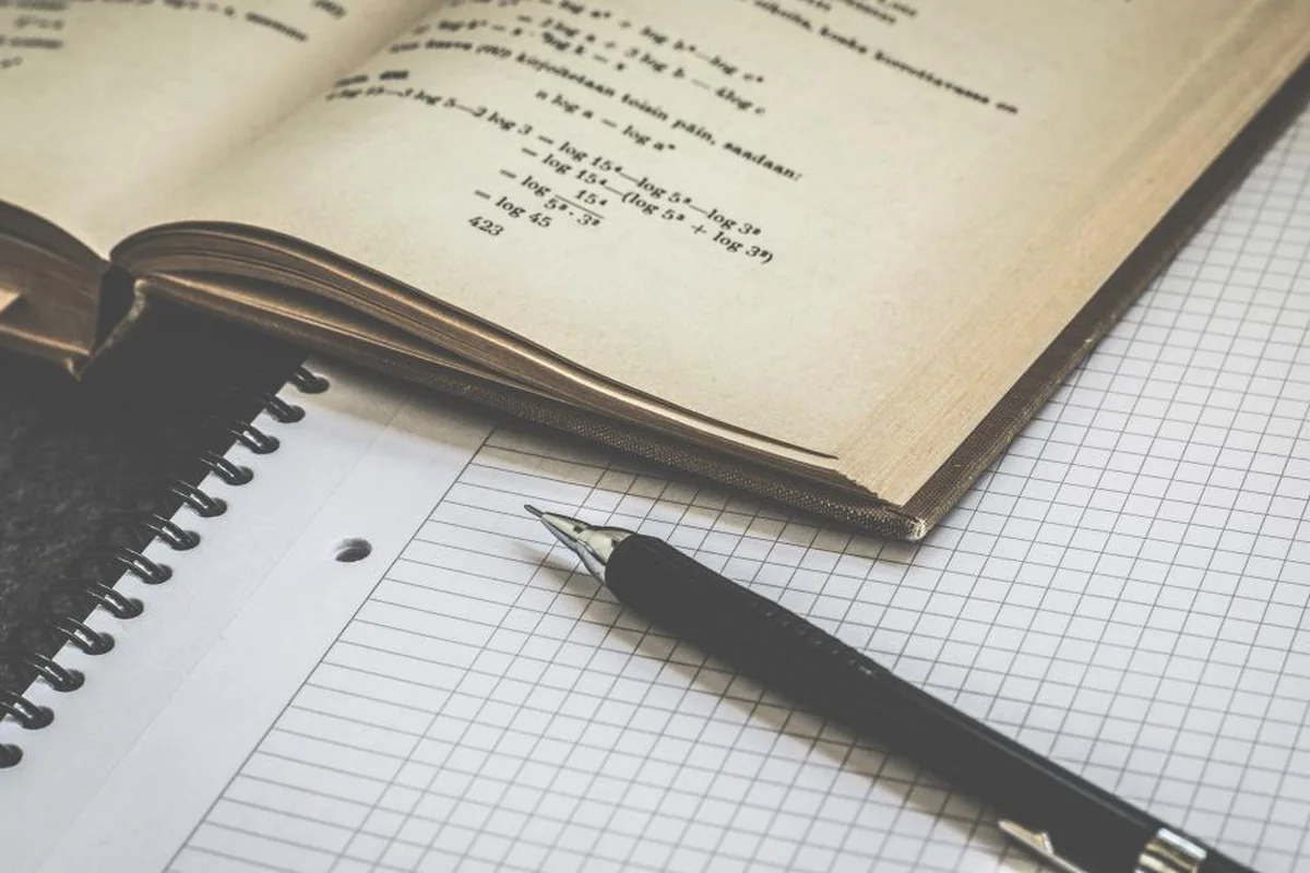 Cuaderno cuadriculado de anillas con una pluma estilográfica junto a un libro abierto con ejercicios de matemáticas