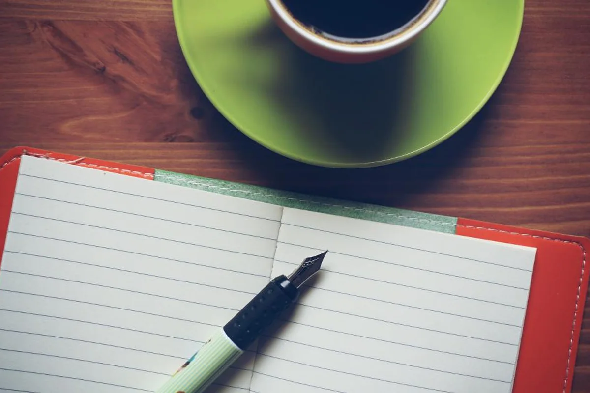 Cuaderno rayado abierto y en blanco con una pluma estilográfica junto a una taza de café sobre una mesa de madera
