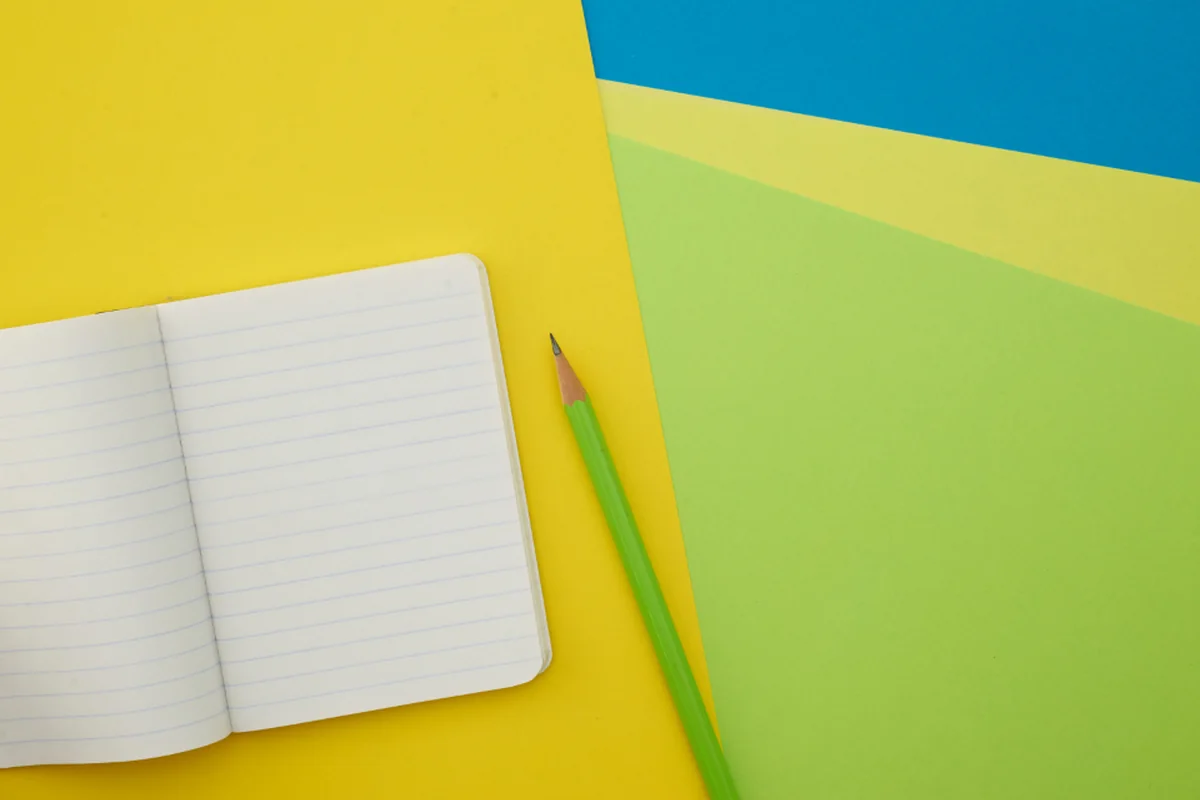 Cuaderno abierto de hojas rayadas y un lápiz verde sobre cartulinas amarillas, verdes y azules