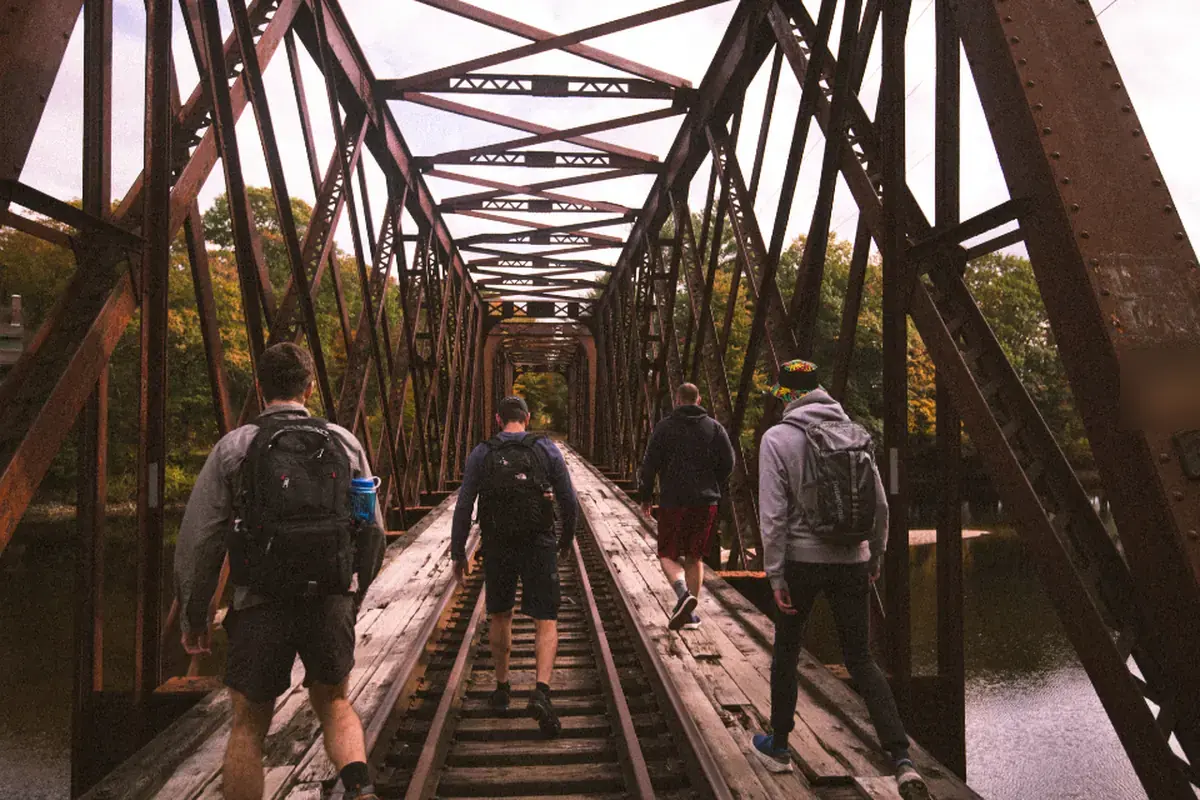 Cuatro jóvenes con mochilas caminando de espaldas por las vías de un viejo puente de hierro rodeado de árboles