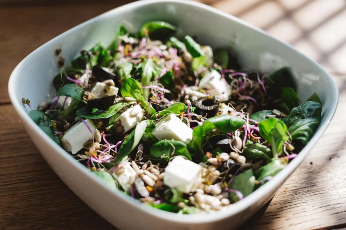 Cuenco blanco con ensalada de hojas verdes, dados de queso fresco, aceitunas, semillas y brotes sobre una mesa de madera iluminada por la luz de una ventana