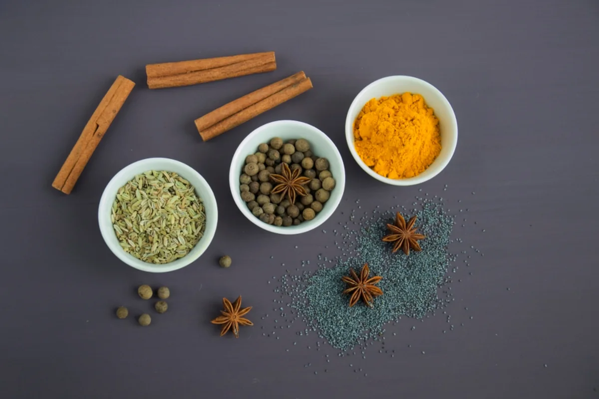 Tres cuencos blancos con especias de colores, ramas de canela y anís estrellado sobre una mesa gris