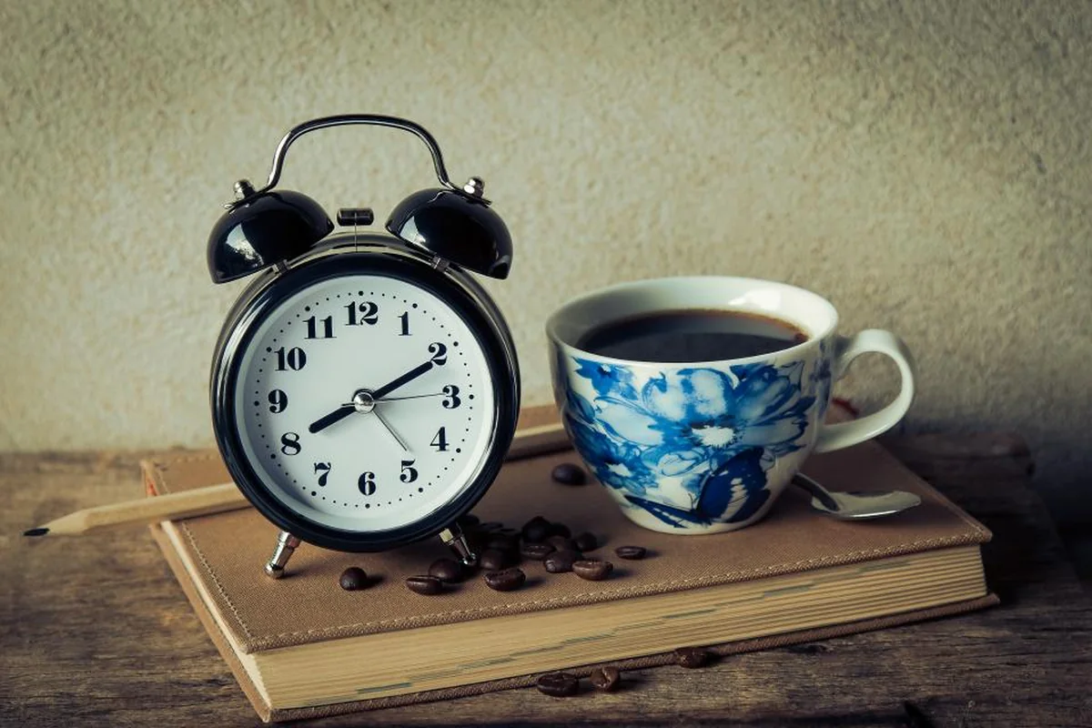 Un despertador negro de campanas y una taza de café con flores azules sobre un libro cerrado, con granos de café esparcidos