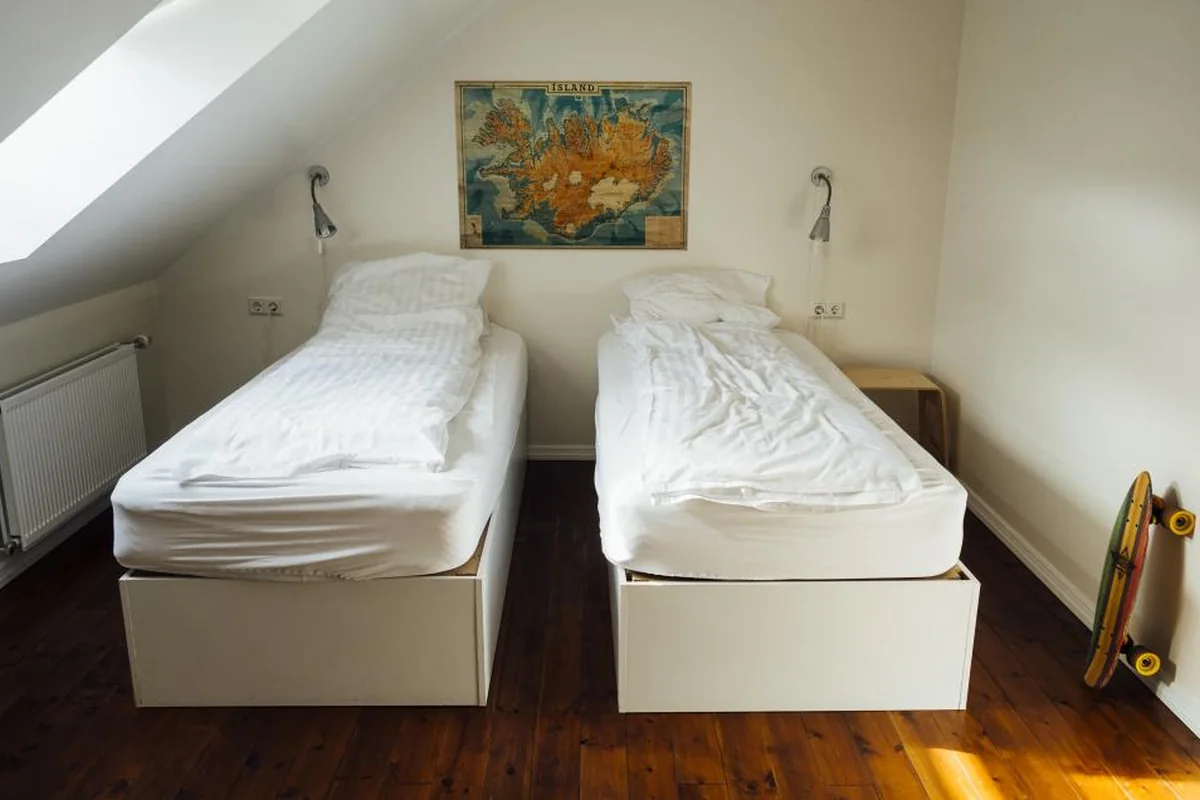 Dormitorio abuhardillado con dos camas individuales blancas, dos lámparas de lectura y un monopatín apoyado en la pared