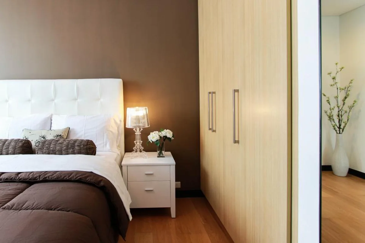 Dormitorio con cama de cabecero blanco, mesilla con una lámpara encendida, un pequeño ramo de flores y un armario de madera clara