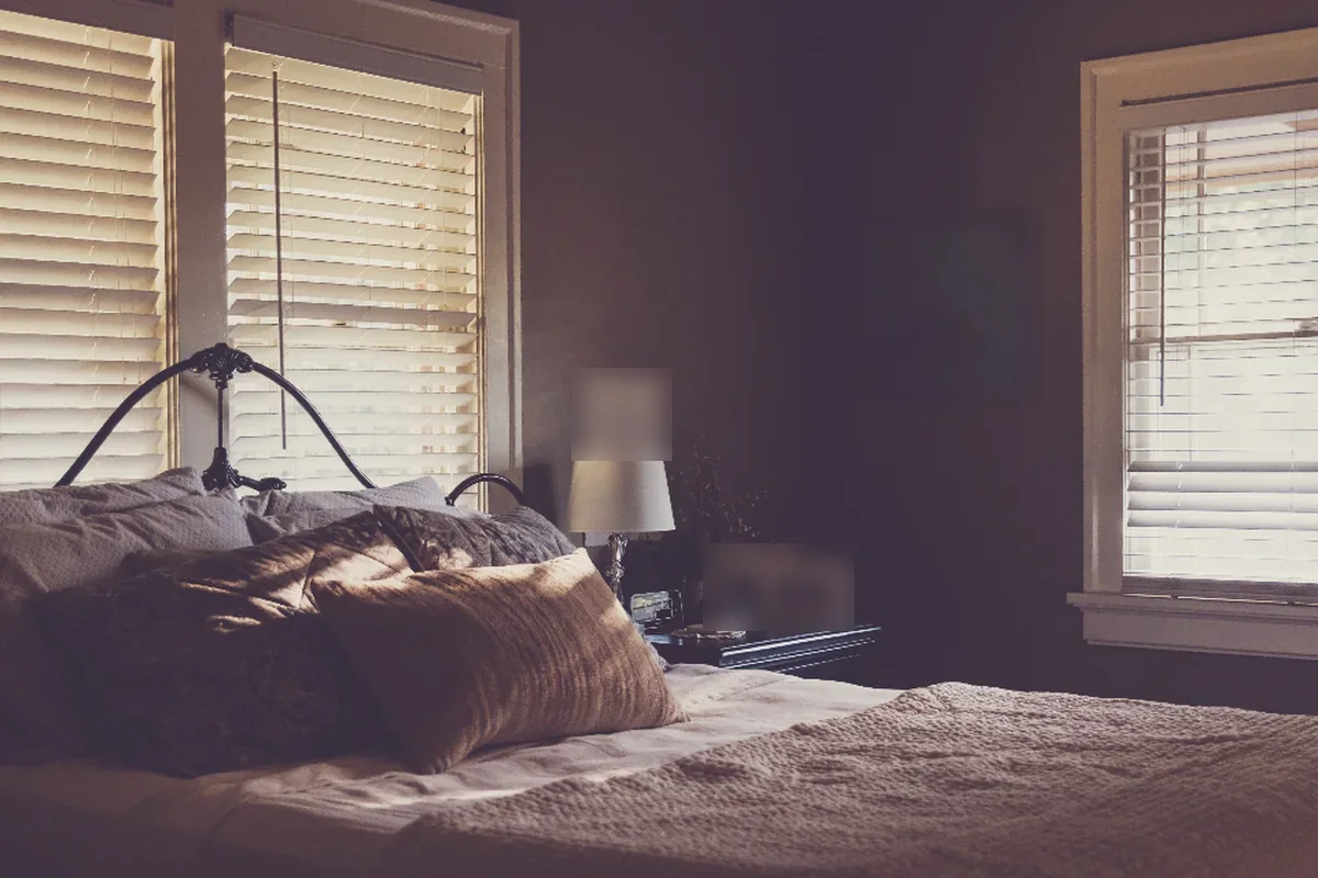 Un dormitorio en penumbra con una cama de cabecero de forja, cojines y luz tenue que entra por las persianas