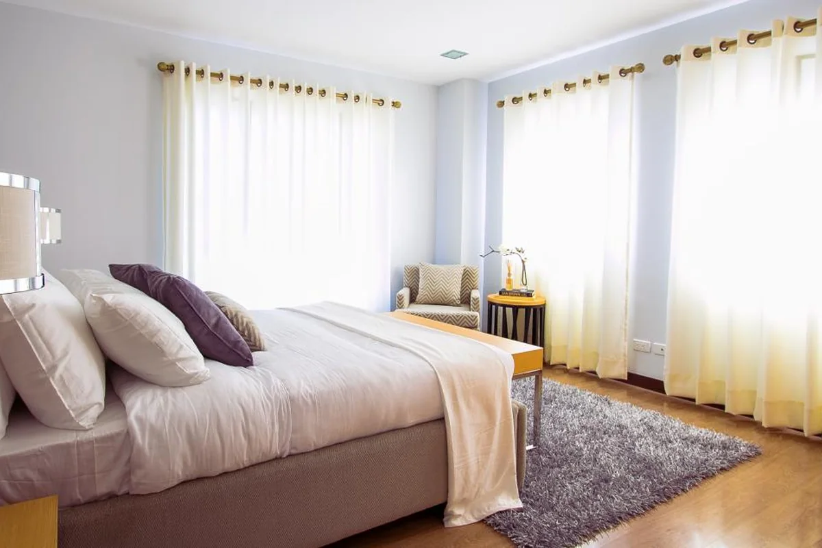 Dormitorio luminoso con una cama doble con cojines morados, una alfombra gris y cortinas claras en las ventanas