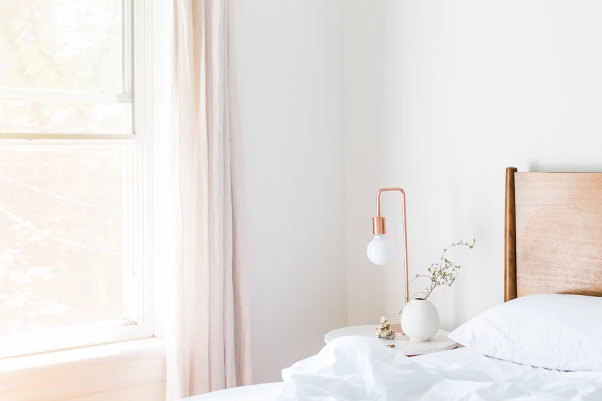 Dormitorio luminoso con una cama de sábanas blancas, una lámpara de mesilla y una ventana con cortina clara