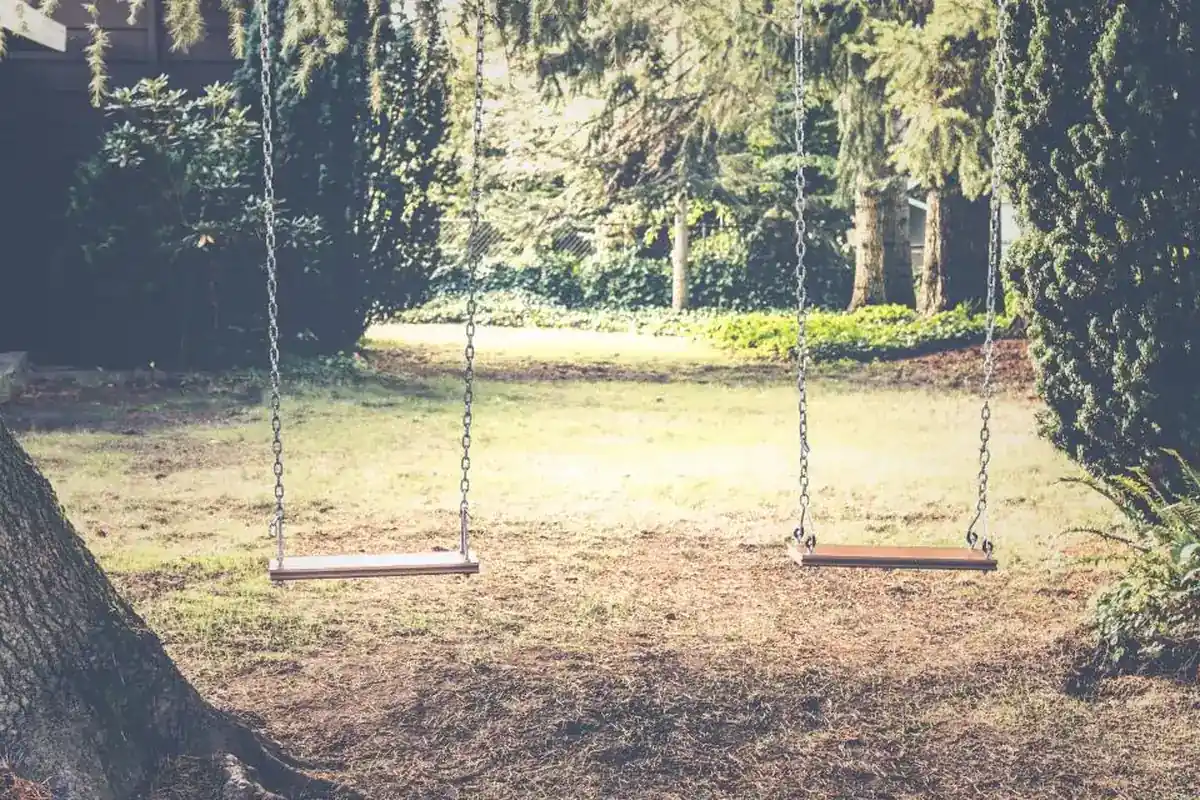 Dos columpios de madera vacíos colgados de cadenas en un jardín con árboles y césped iluminado por el sol