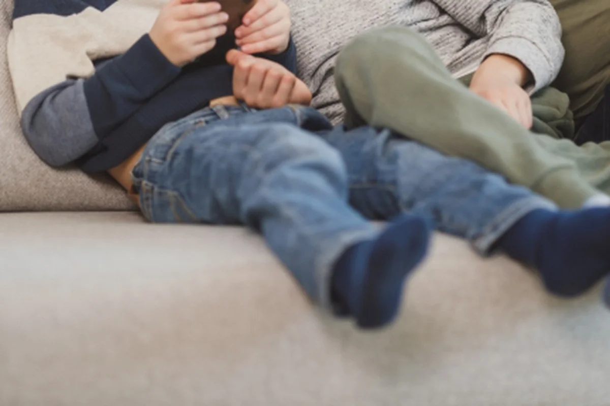 Dos niños sentados juntos en un sofá gris, vistos de cintura para abajo, con vaqueros y calcetines azules