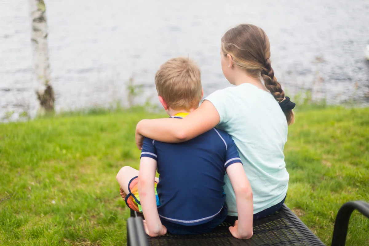 Dos niños de espaldas sentados en un banco frente a un lago; la mayor rodea con el brazo al pequeño