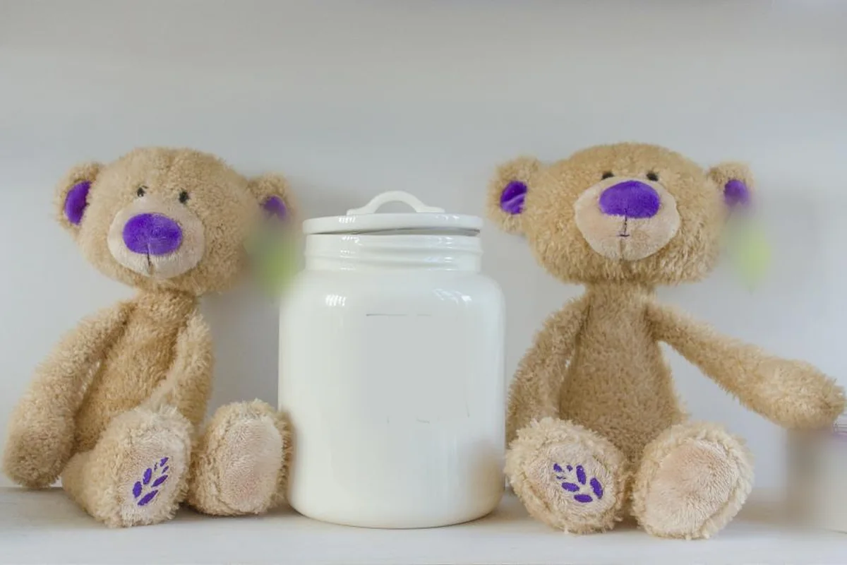 Dos ositos de peluche de color crema con orejas y nariz moradas sentados en una balda, uno a cada lado de un tarro blanco de cerámica