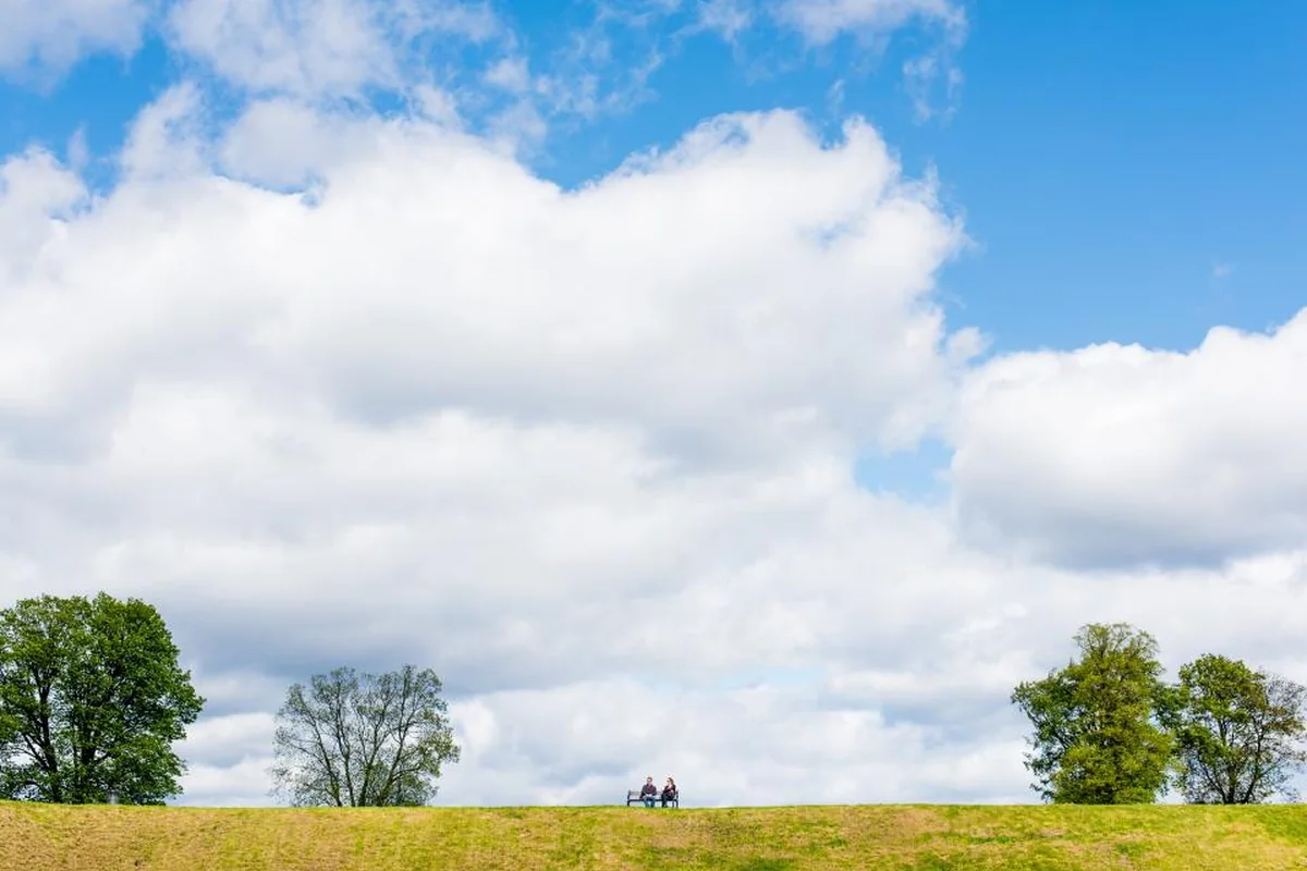 Dos personas sentadas en un banco a lo lejos, en lo alto de una colina verde, bajo un cielo azul con grandes nubes blancas
