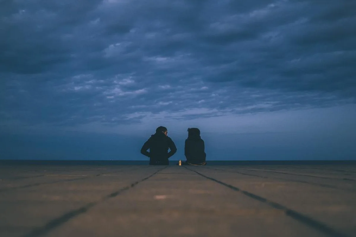 Dos personas sentadas una junto a otra de espaldas, contemplando el horizonte bajo un cielo azul nublado al anochecer