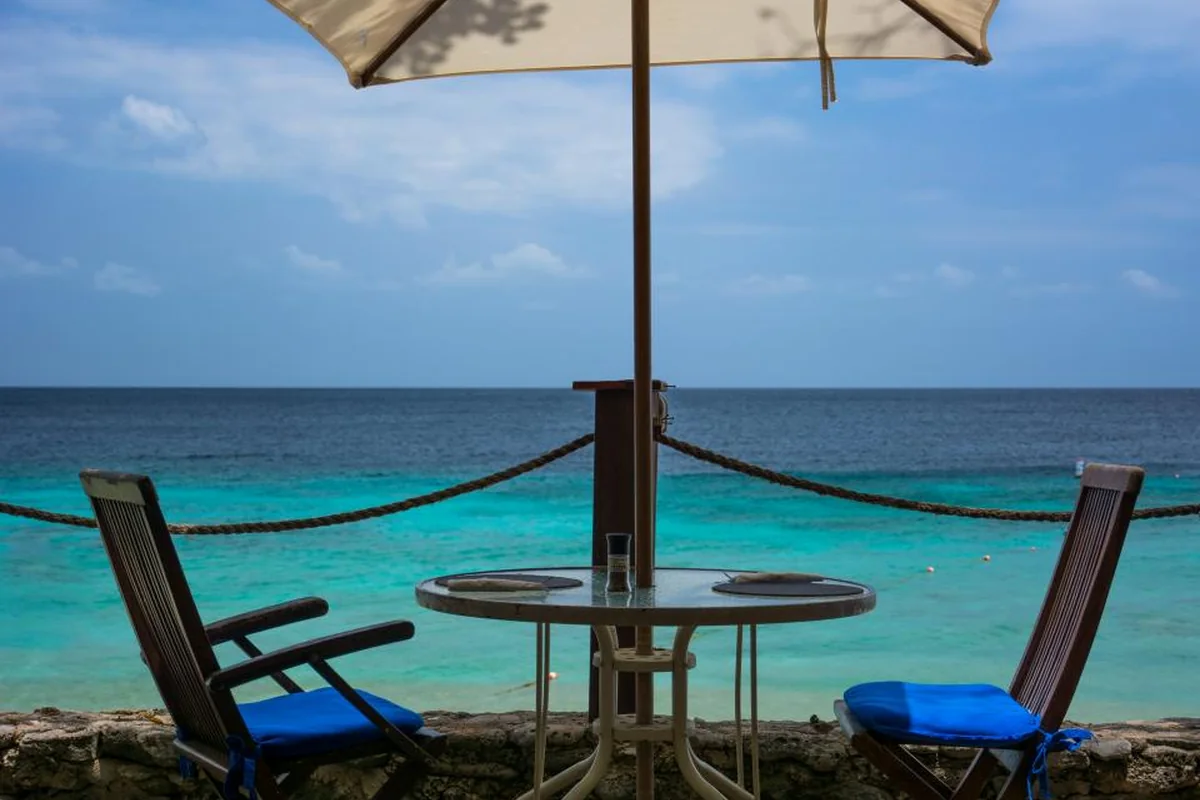 Dos sillas de madera con cojines azules y una mesa redonda bajo una sombrilla, frente al mar
