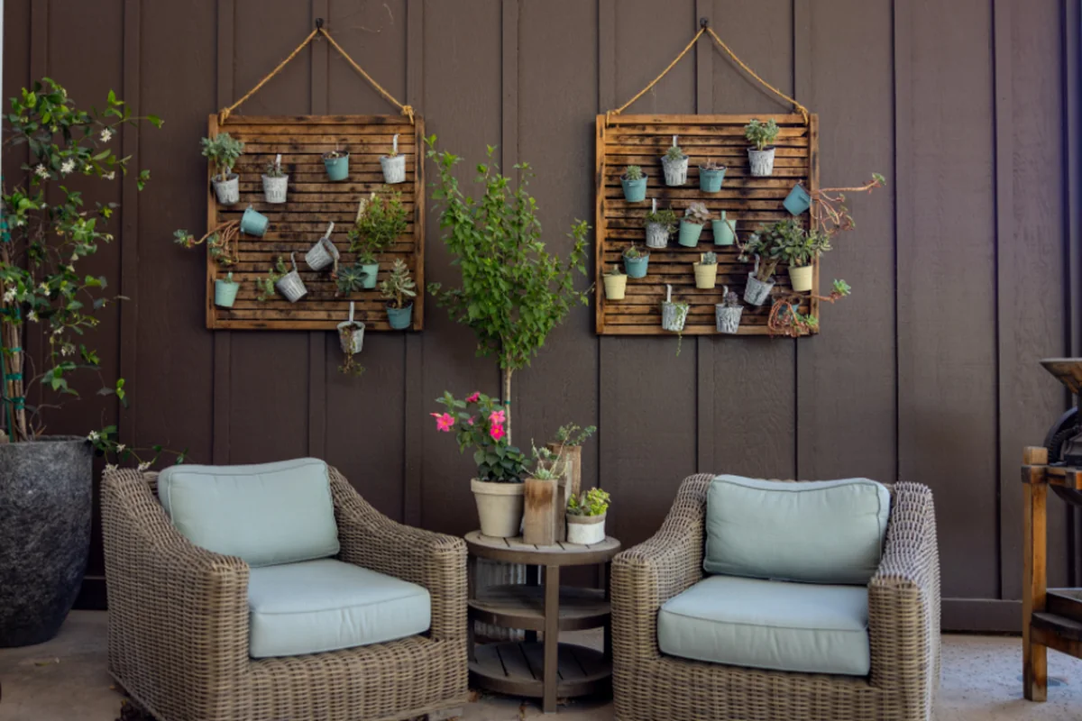 Dos sillones de mimbre con cojines claros, uno frente a otro, junto a una mesita con plantas en un patio con pared de madera oscura