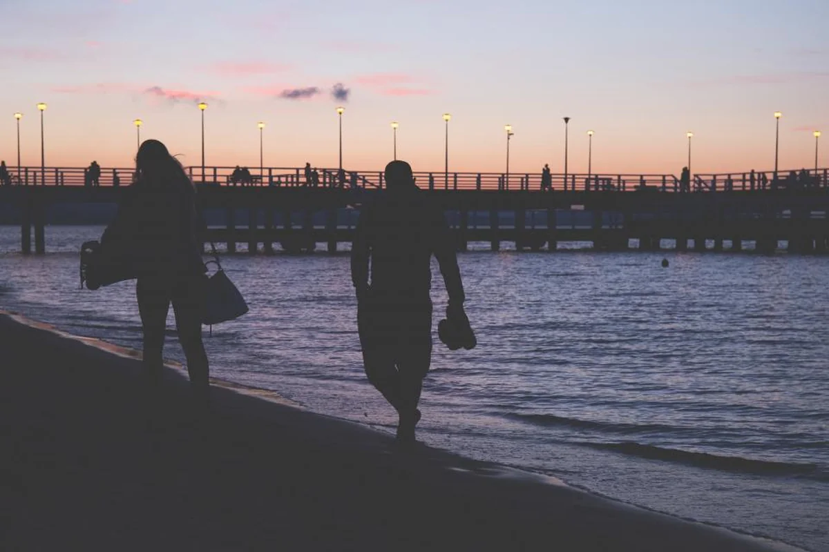 Dos siluetas caminando por la orilla del mar al anochecer, separadas por unos metros, con un muelle iluminado al fondo