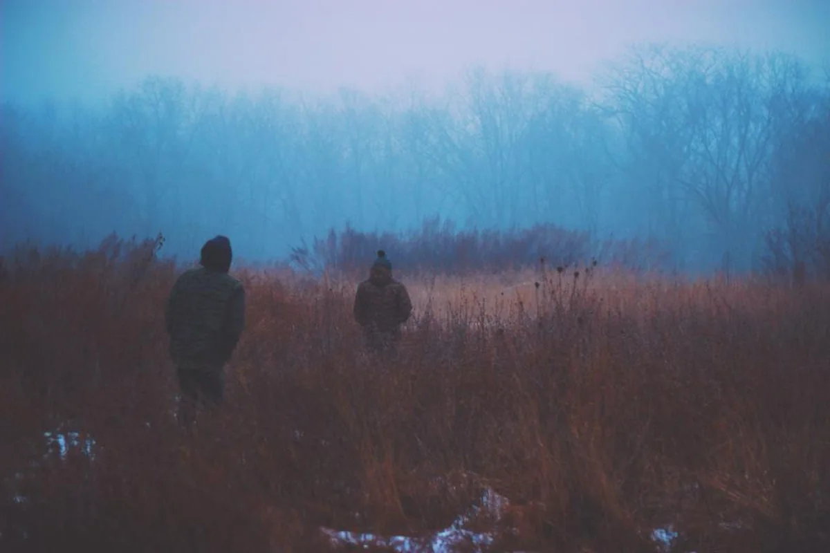 Dos personas con gorro de lana, de espaldas y desenfocadas, entre hierba alta en un campo cubierto de niebla azulada
