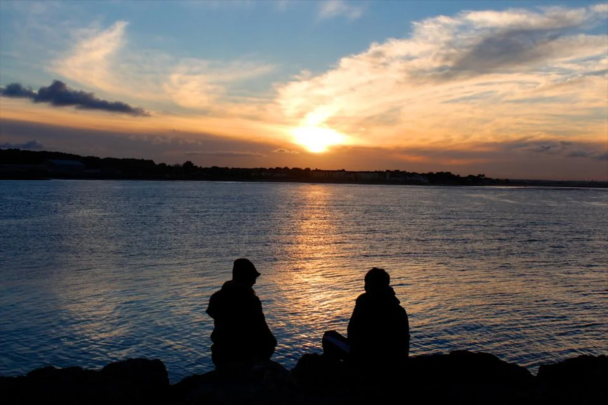 Dos siluetas sentadas sobre unas rocas frente al mar al atardecer, separadas por un pequeño espacio