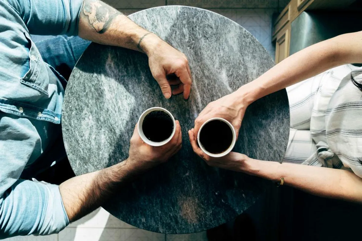 Vista cenital de dos personas sentadas frente a frente en una mesa redonda de mármol gris, cada una sosteniendo una taza de café solo