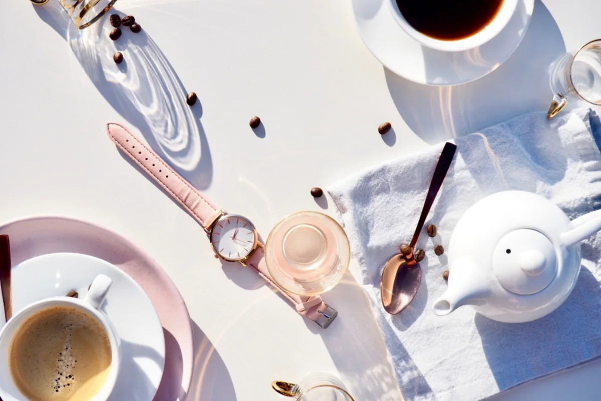 Vista cenital de una mesa blanca con dos tazas de café, una tetera, un reloj de correa rosa, una cuchara dorada y granos de café sueltos