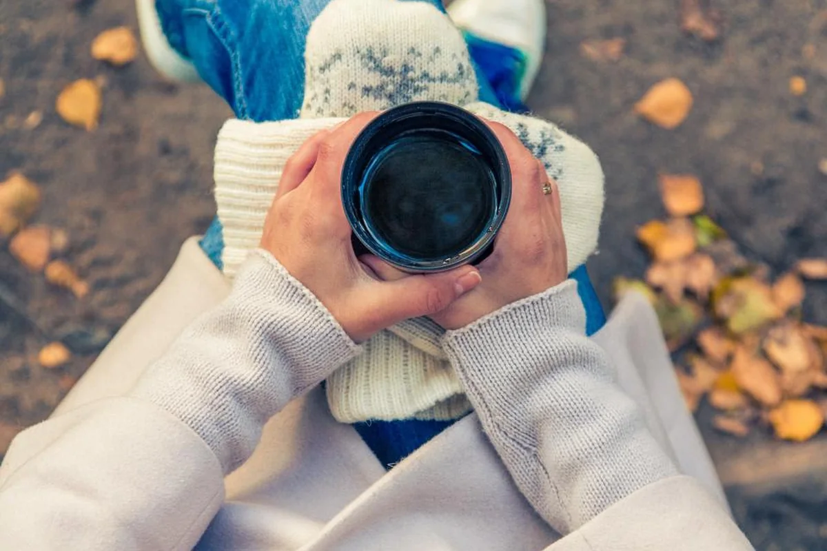 Manos sujetando una taza caliente, envueltas en las mangas de un jersey, al aire libre