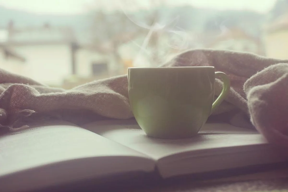 Taza humeante sobre un libro abierto, junto a una manta, en un ambiente cálido