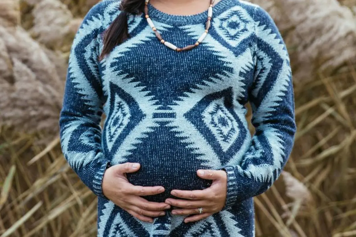 Mujer embarazada con jersey azul de rombos apoyando las manos en el vientre, en un campo de hierba seca