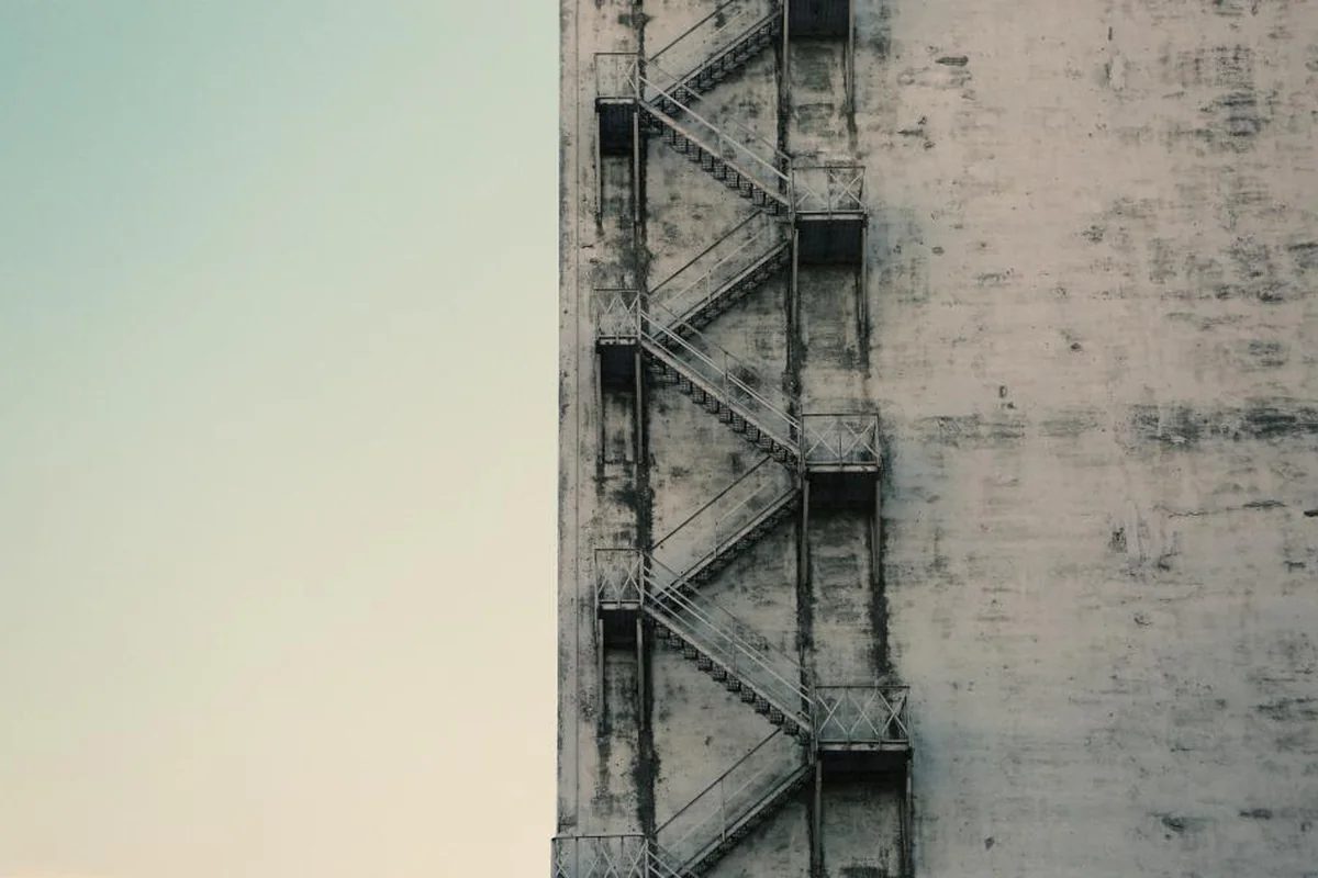 Una escalera metálica exterior en zigzag adosada a una fachada de hormigón gris, bajo un cielo claro