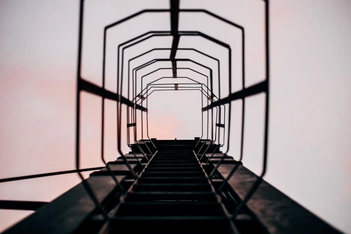 Escalera metálica con aros de protección vista desde abajo, ascendiendo hacia un cielo claro de tonos rosados