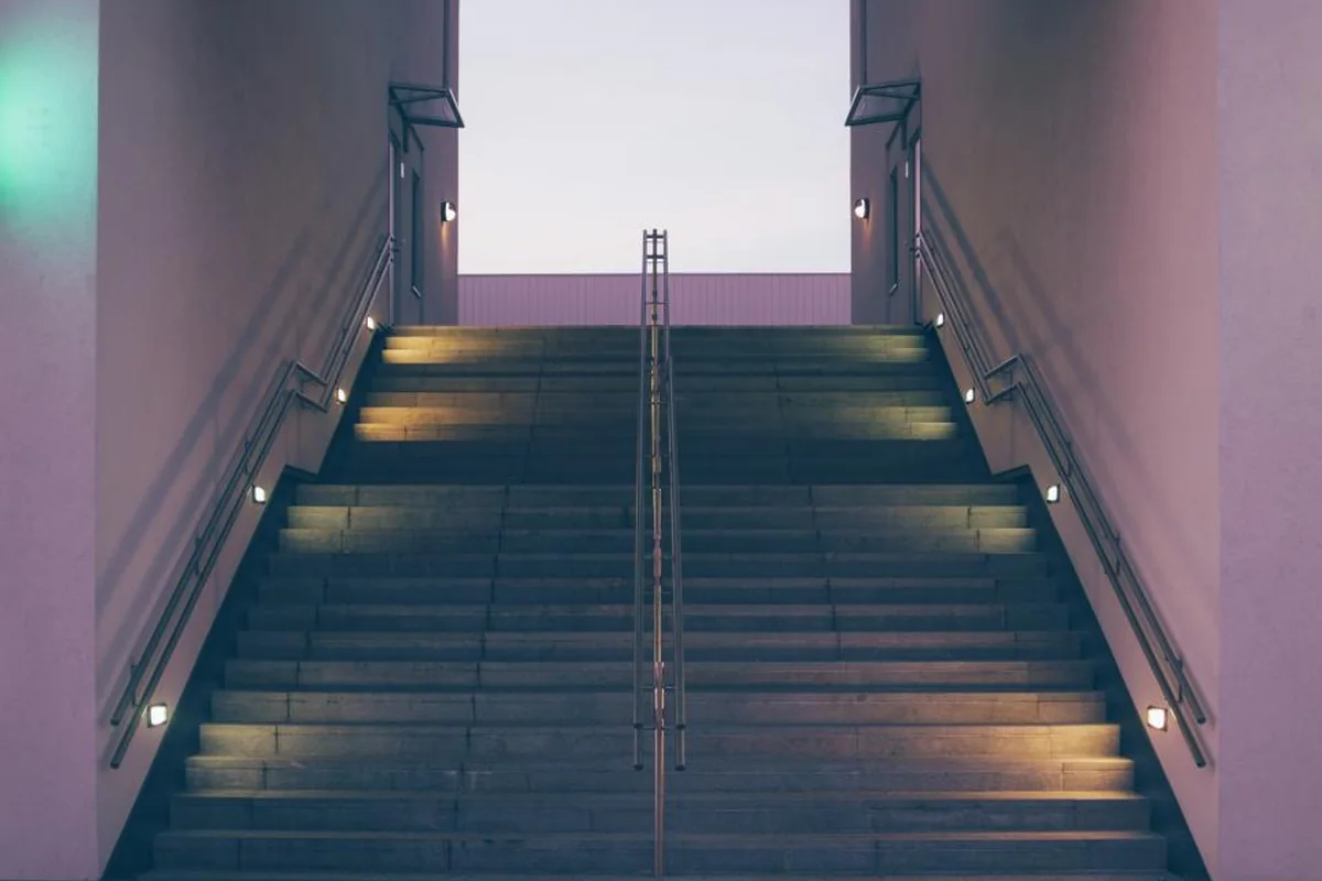 Escalera ancha iluminada por pequeñas luces laterales que sube entre dos paredes rosadas hacia un cielo claro