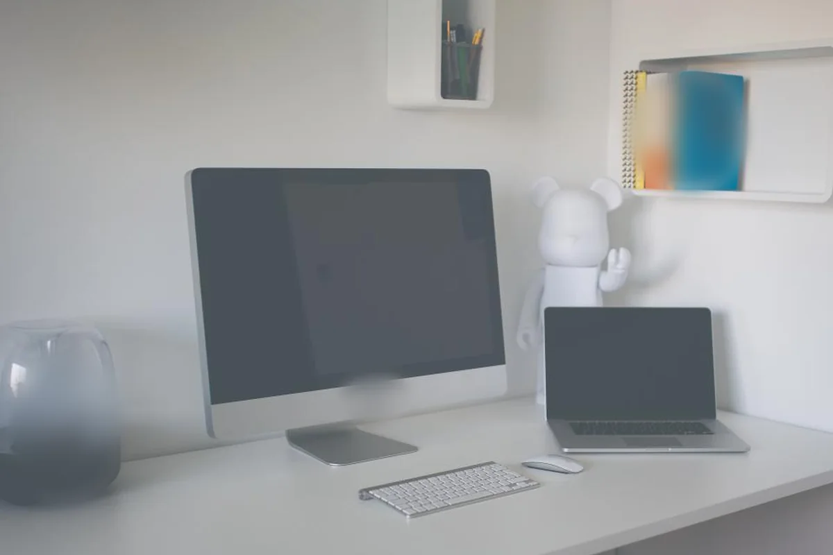 Escritorio blanco con un ordenador de sobremesa y un portátil apagados, un teclado, un ratón y una figura blanca decorativa junto a una estantería con libros