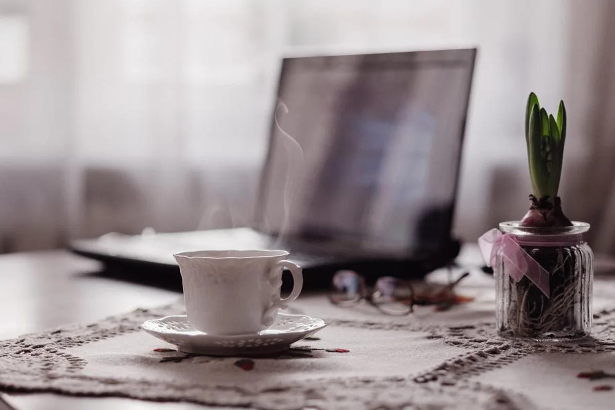 Portátil abierto y una taza de café humeante sobre una mesa junto a la ventana