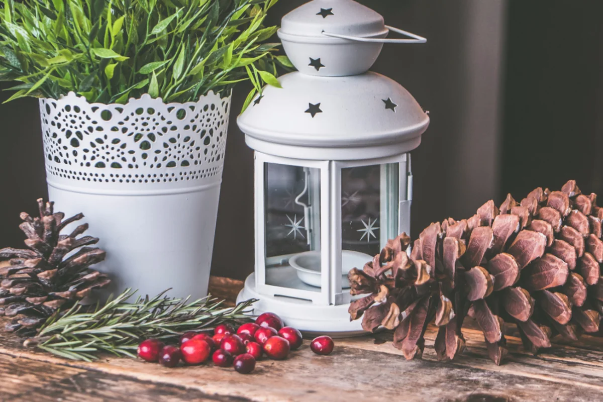 Farol blanco con estrellas caladas junto a una maceta, piñas, ramitas de romero y arándanos rojos sobre una mesa de madera