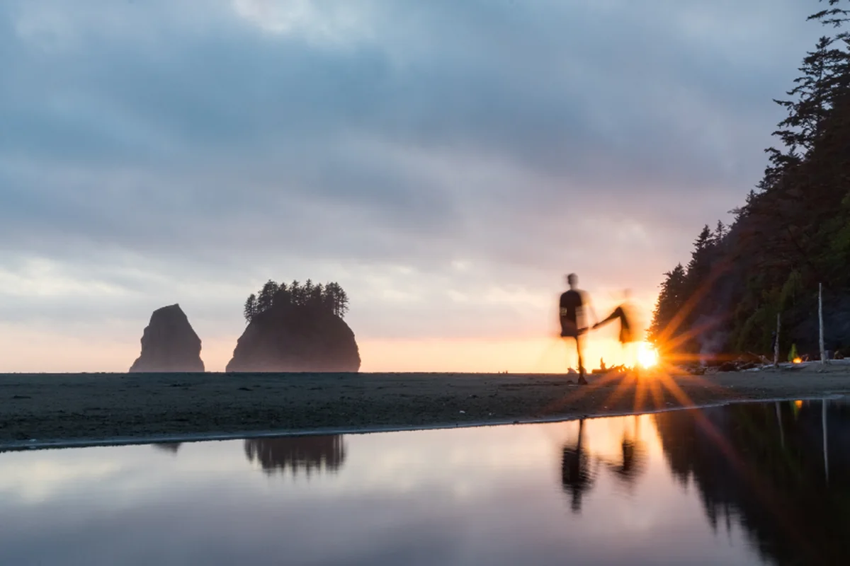 Dos figuras desenfocadas caminando por la orilla de una playa al atardecer, con el sol entre los árboles y dos islotes reflejados en el agua