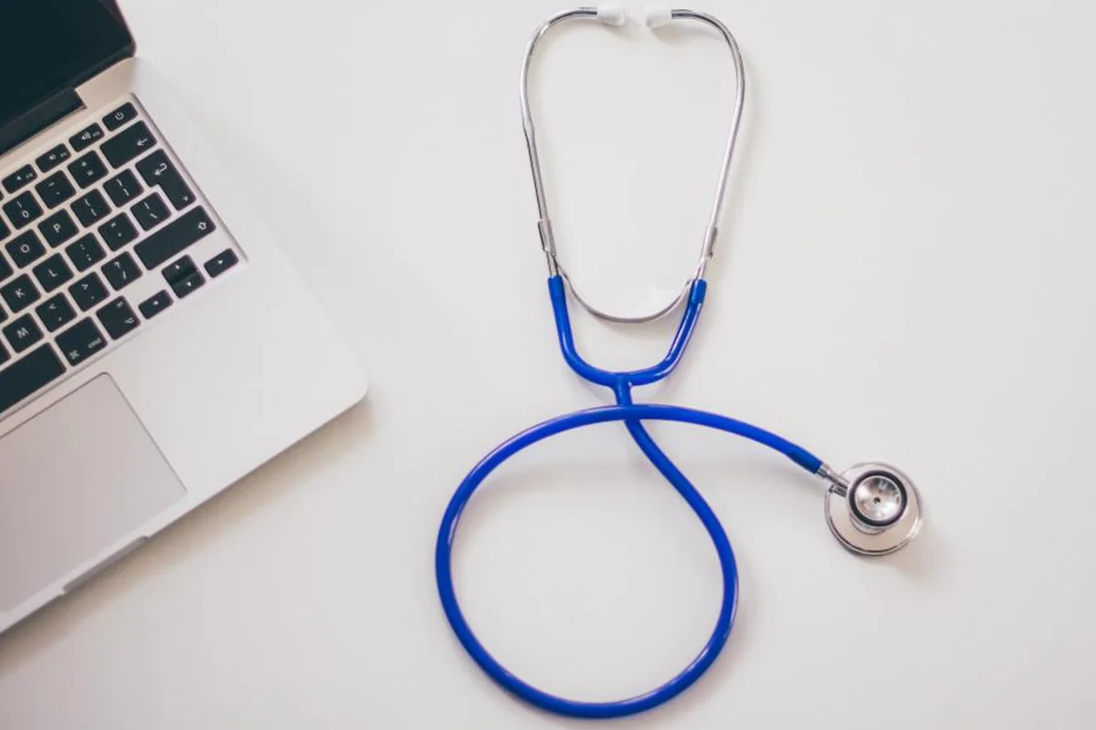 Fonendoscopio azul enrollado sobre una mesa blanca junto a un ordenador portátil abierto