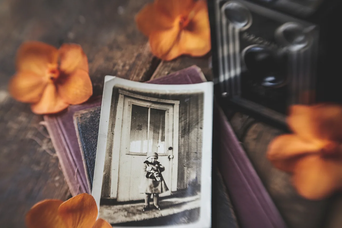 Fotografía antigua en blanco y negro de un niño ante una puerta, sobre un libro viejo rodeado de flores naranjas y una cámara antigua