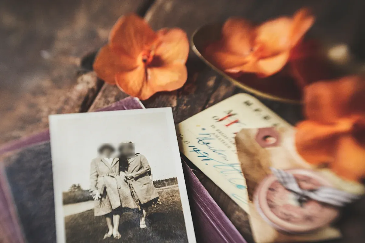 Fotografía antigua en blanco y negro de dos personas abrigadas, sobre un libro y junto a flores naranjas y una etiqueta escrita a mano