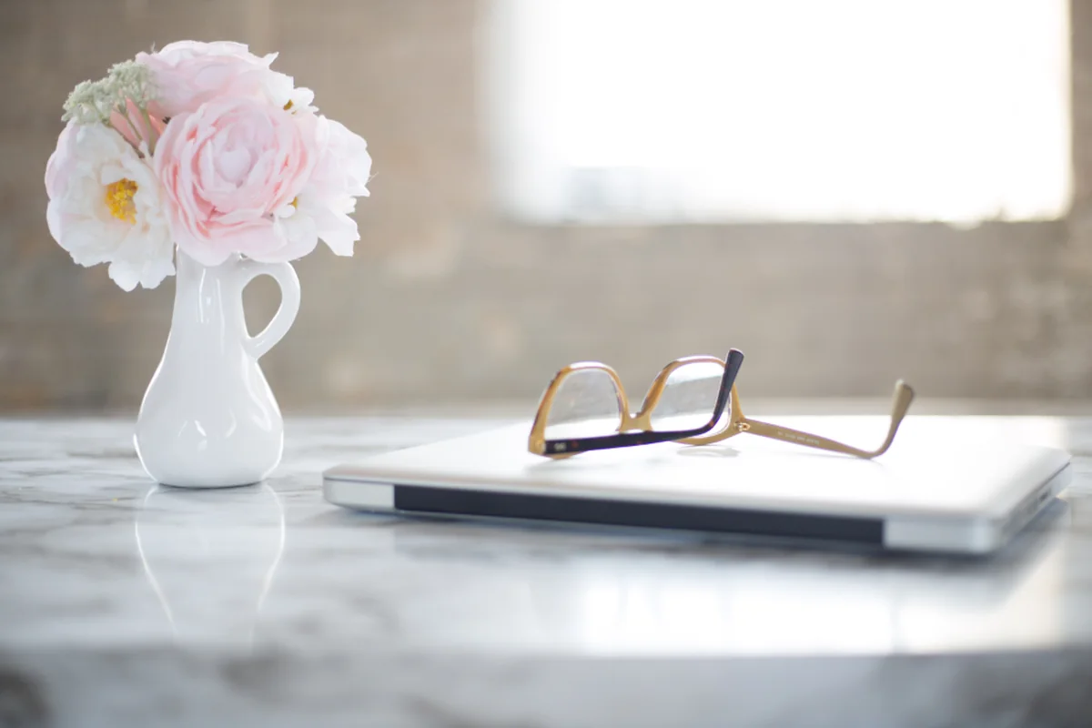 Unas gafas de montura dorada apoyadas sobre un portátil cerrado, junto a un pequeño jarrón blanco con flores rosas, frente a una ventana luminosa