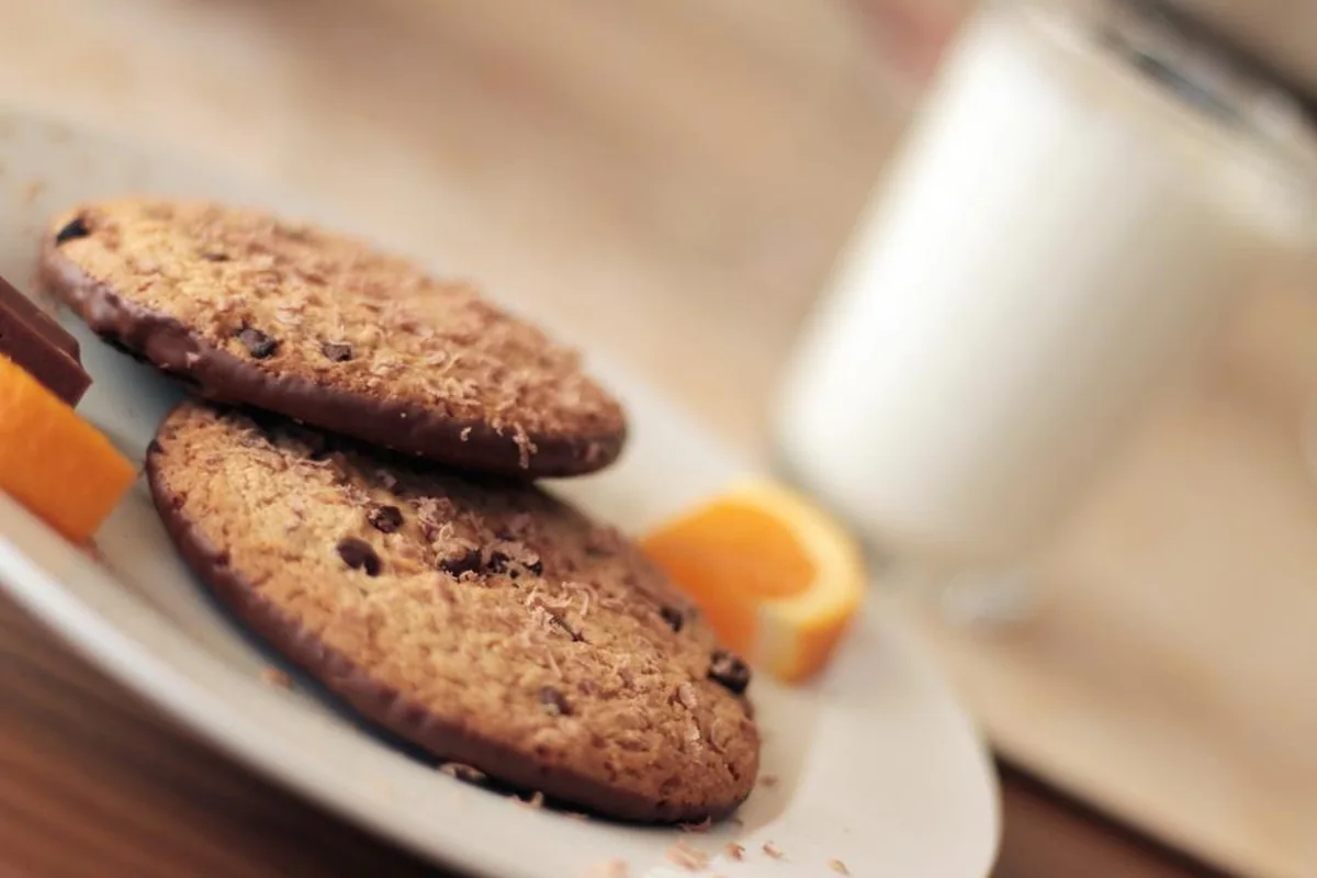 Dos galletas con pepitas de chocolate en un plato junto a gajos de naranja y un vaso de leche desenfocado