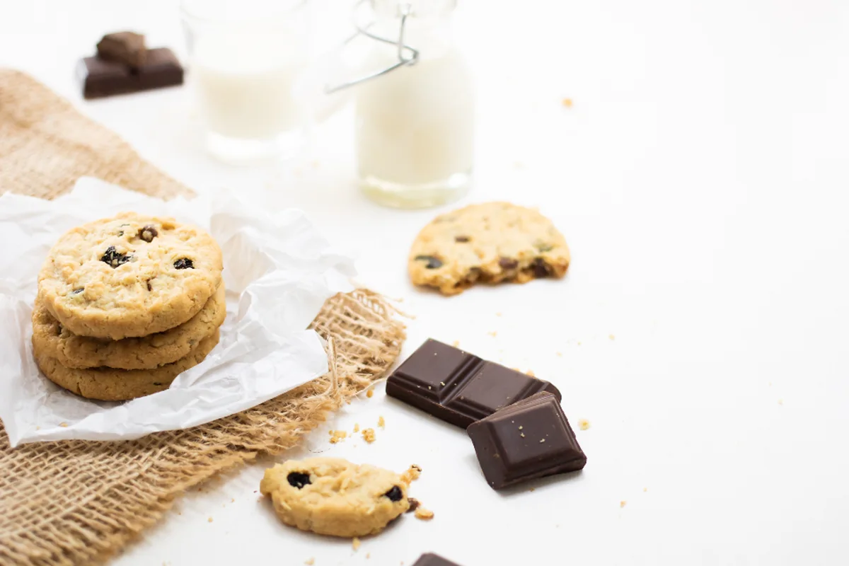 Galletas con pepitas, una de ellas mordida, y onzas de chocolate sobre una mesa blanca junto a dos botellas de leche