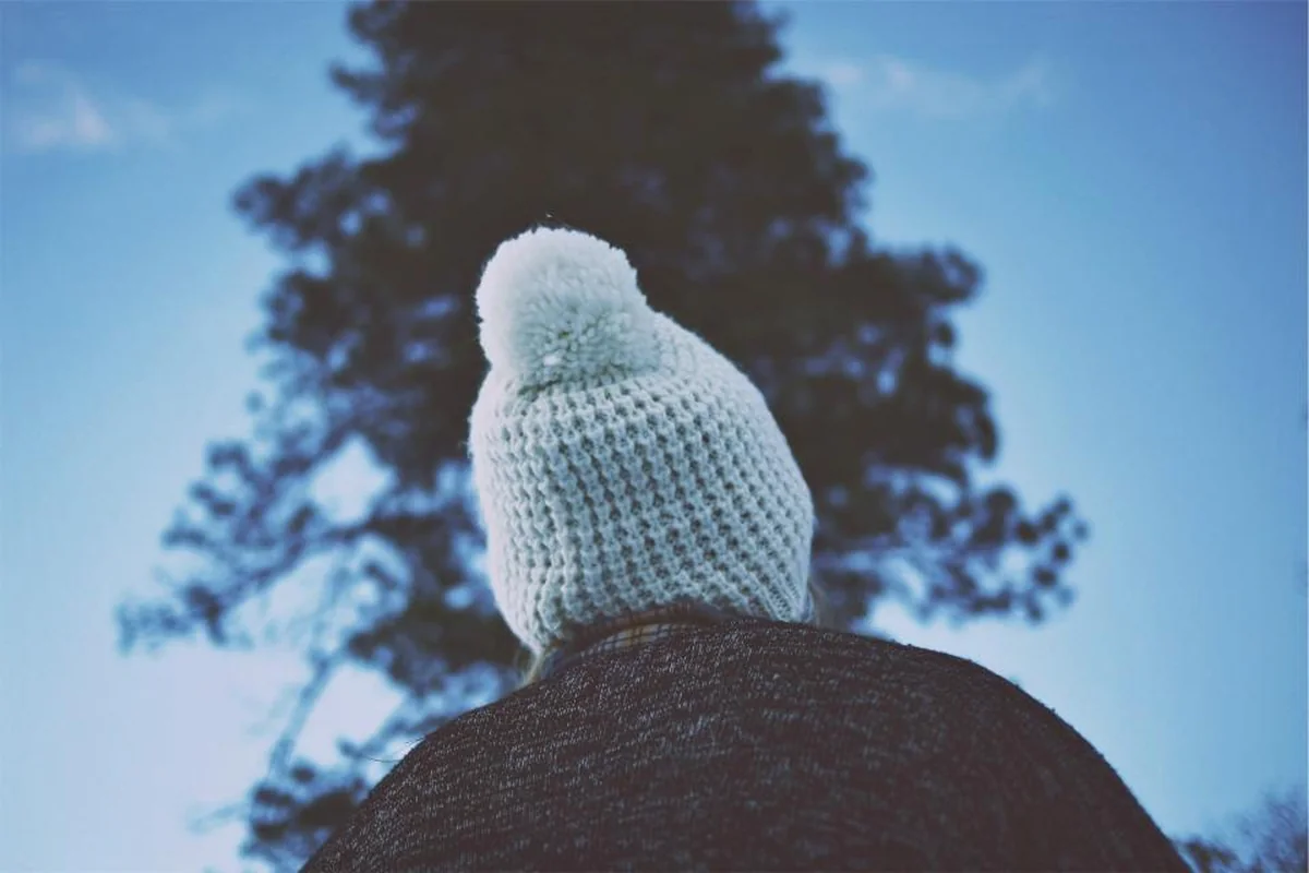 Cabeza de una persona vista desde atrás con un gorro de lana claro, recortada contra un cielo azul y la copa oscura de un árbol