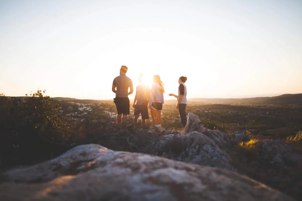 Cuatro personas de pie y de espaldas sobre unas rocas en lo alto de una colina, recortadas a contraluz frente al sol del atardecer