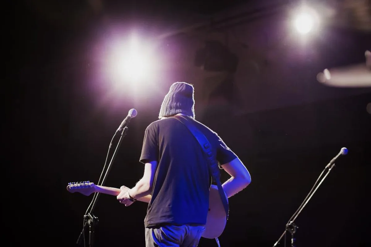 Guitarrista de espaldas en un escenario oscuro, iluminado por focos de luz blanca y violeta, con dos micrófonos en sus pies