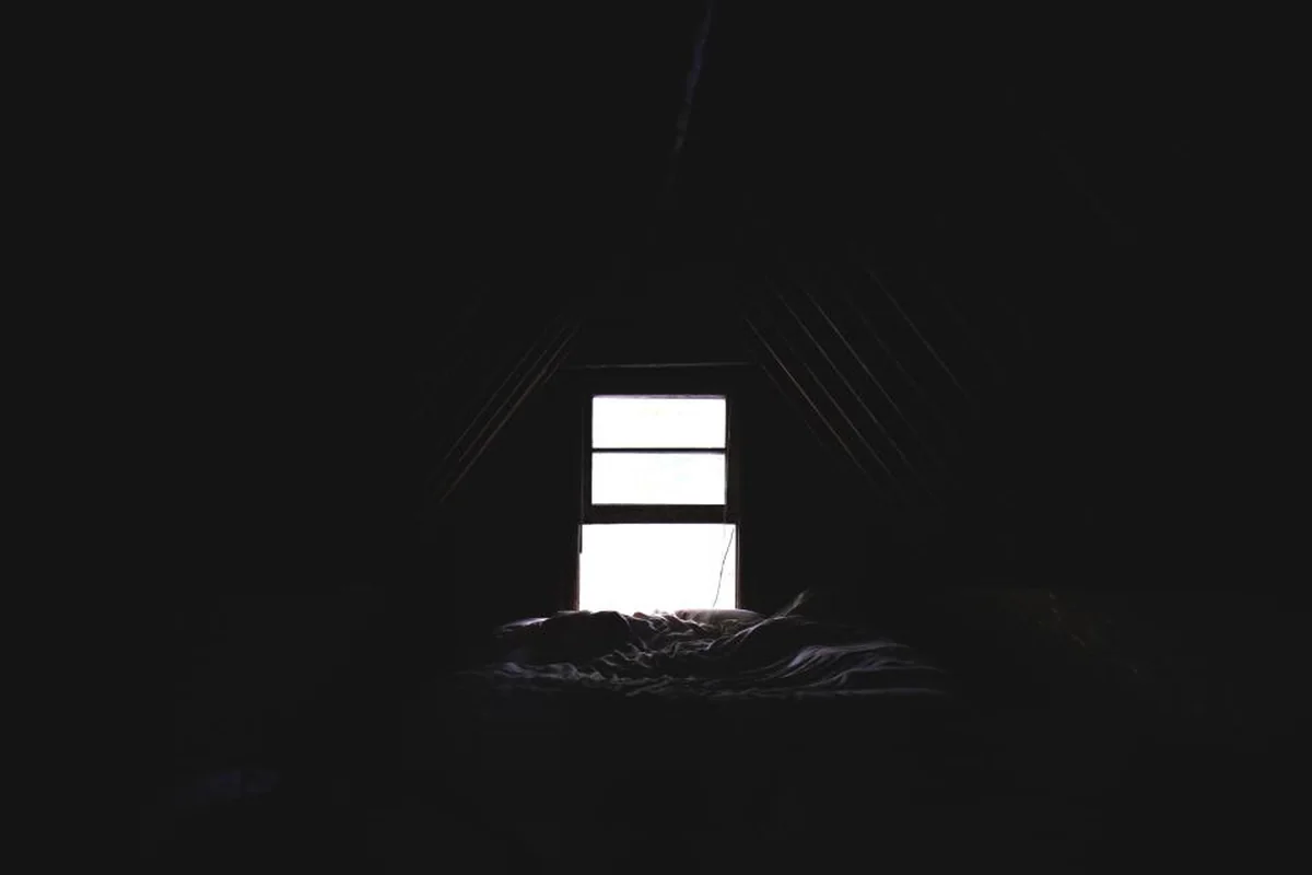Una habitación abuhardillada casi a oscuras con una cama deshecha bajo una ventana pequeña por la que entra luz blanca