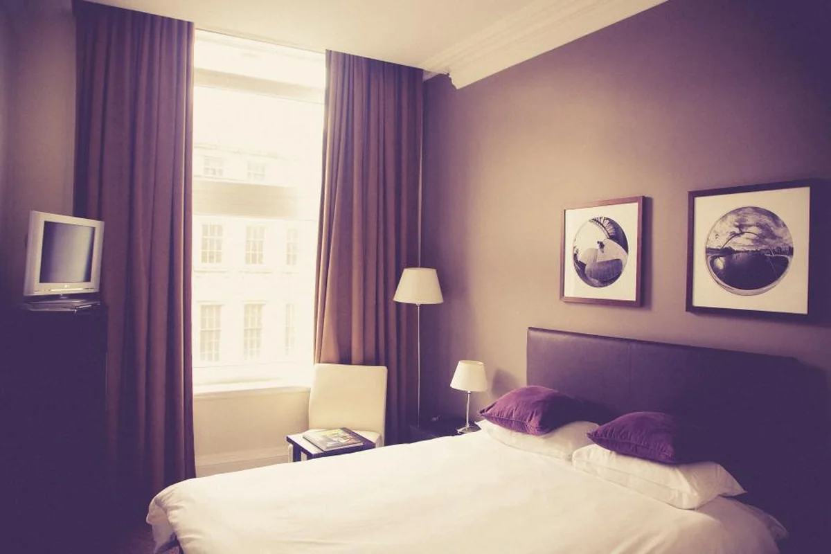 Una habitación con una cama de sábanas blancas y cojines morados, dos cuadros en la pared, una lámpara encendida y una ventana luminosa con cortinas moradas
