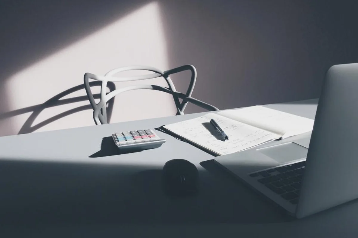 Una habitación en penumbra con un escritorio, un portátil abierto, un cuaderno y una silla vacía iluminada por una franja de luz