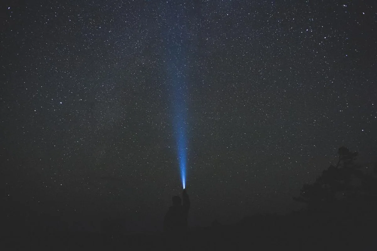 Un haz de luz de linterna dirigido hacia el cielo estrellado en una noche oscura con algo de niebla, junto a una silueta apenas visible