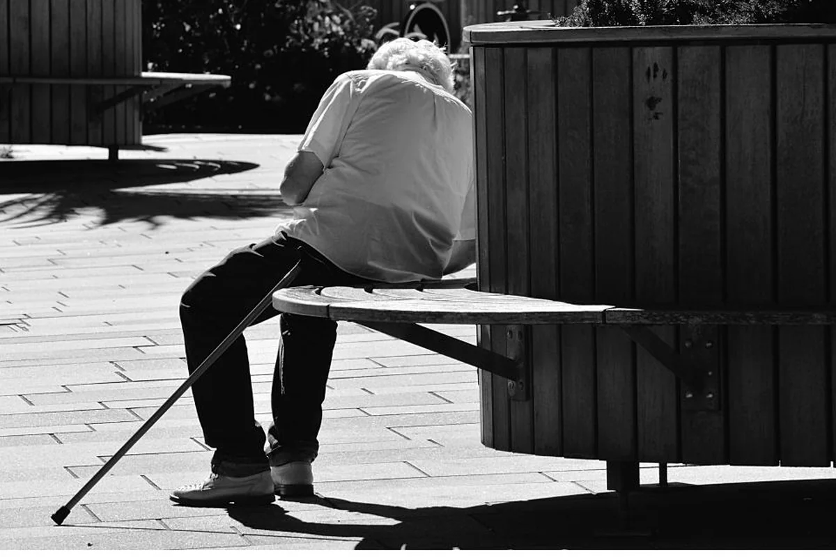 Hombre mayor de espaldas sentado en un banco de madera con un bastón apoyado en el suelo, en blanco y negro